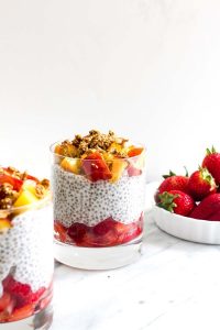 A cup filled with sliced strawberries then chia seed pudding then chopped peaches with granola with a side bowl of strawberries.