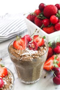 A glass of chocolate chia seed pudding topped with berries and being sprinkled with coconut.