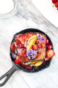 A top view of a cast iron skillet with a stack of french toast with strawberry sauce, flowers, powdered sugar, and peaches.