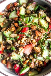 A closeup of a bowl of summer lentil salad with lentils, cucumbers, strawberries, and mint.