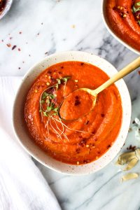 A bowl of spicy roasted red pepper soup garnished with red pepper flakes and micro greens with a spoon taking a bite on a marble background.