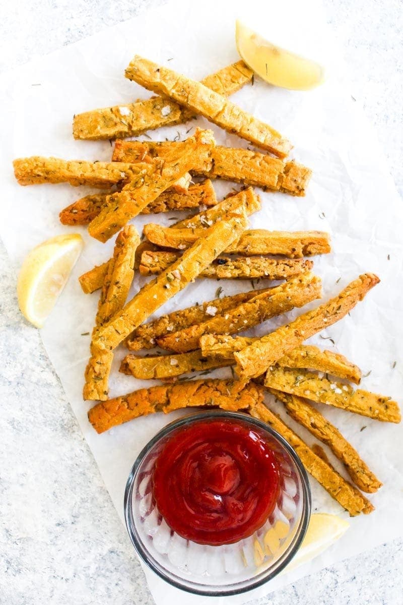 Chickpea fries on parchment paper with lemon slices, salt and a side of ketchup.