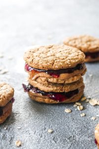 A stack of peanut butter and jelly sandwich cookies on a grey background with rolled oats scattered as a garnish.