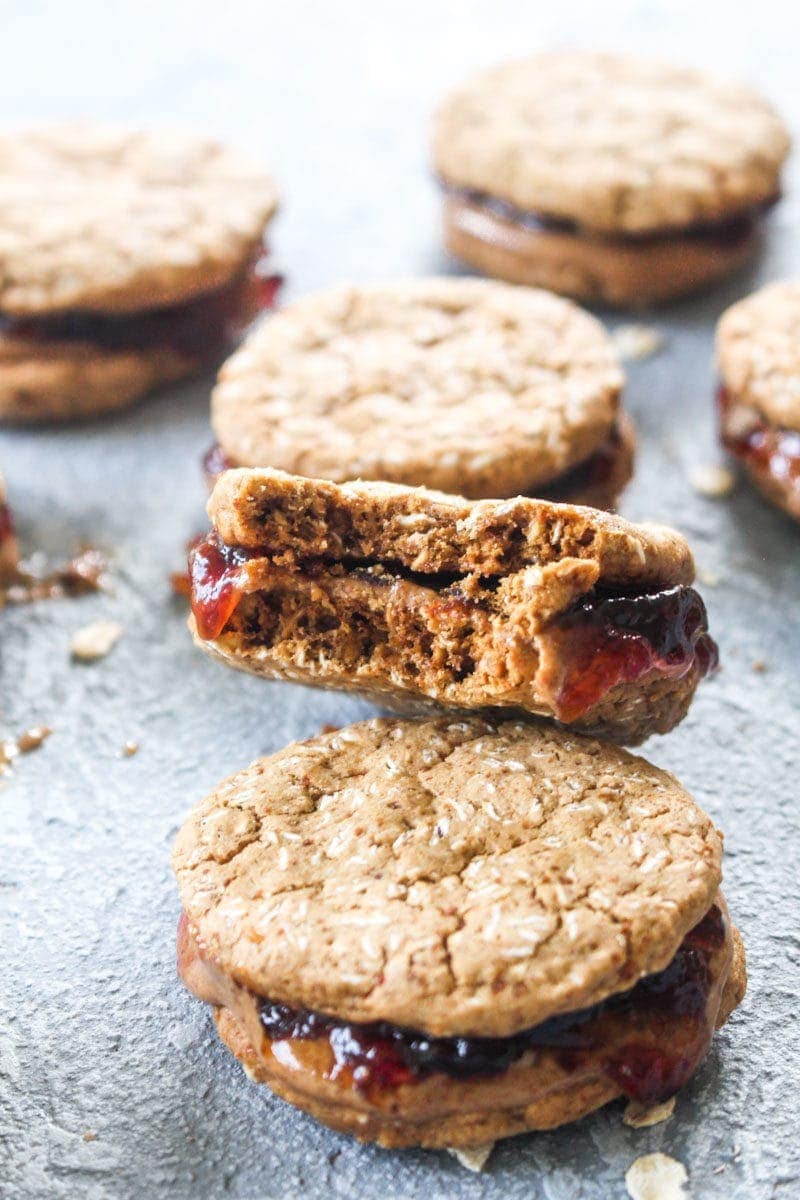 6 peanut butter and jelly sandwich cookies, one with a bite out of it, with raspberry jam, peanut butter, and oats to garnish.