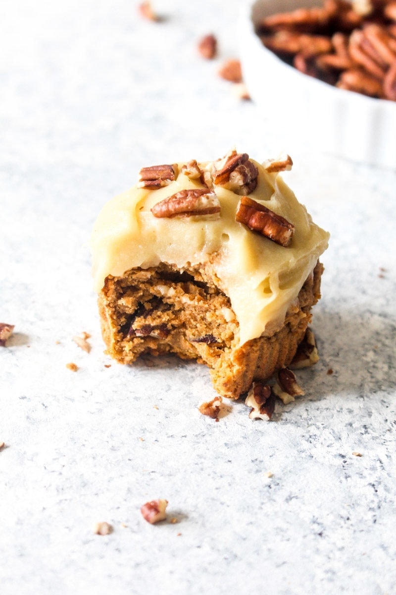 One sweet potato cupcake with sweet potato frosting and a bowl of pecans on a white background.