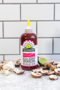 A bottle of Blue Agave Sriracha by Yellowbird Foods with sliced mushrooms, lime, garlic, and ginger.