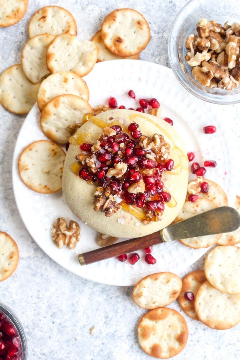 Sharp vegan cheese ball on a white plate topped with walnuts, pomegranates, and walnuts with crackers and a cheese knife.