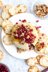 Sharp vegan cheese ball with a slice out of it on a white plate topped with walnuts, pomegranates, and walnuts with crackers.