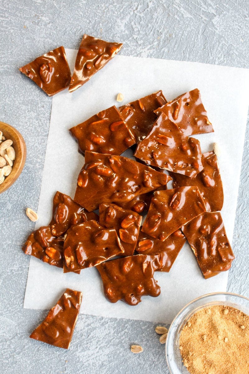 A pile of healthy peanut brittle on a piece of parchment paper with peanuts and coconut sugar in bowls.