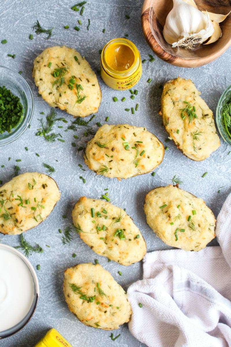 A bunch of creamy mustard twice baked potatoes on a grey background with bowls of herbs and a jar of mustard.