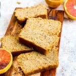 Slices of gluten free orange poppy seed bread on a wood cutting board with halved oranges.