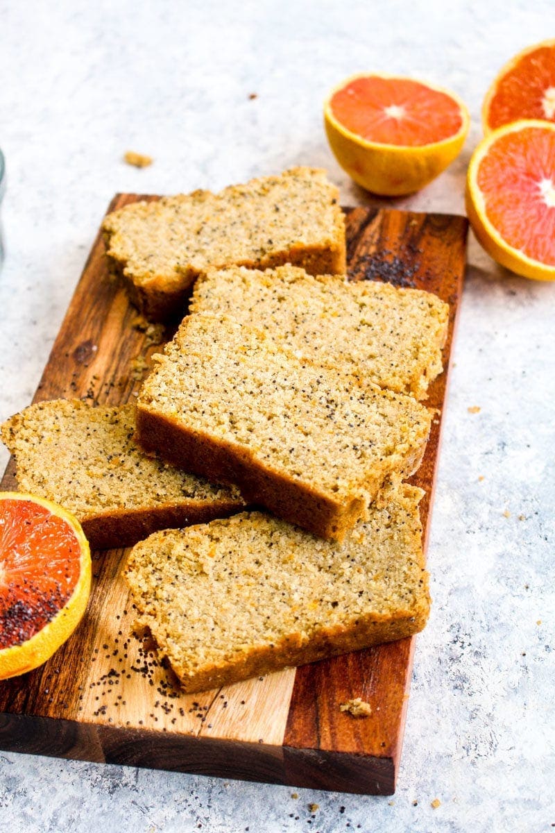 Slices of gluten free orange poppy seed bread on a wood cutting board with halved oranges.
