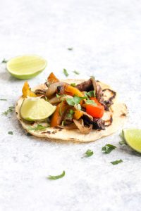 A vegan fajita with bell peppers, portobello mushrooms, and onions on a tortilla with cilantro and lime.