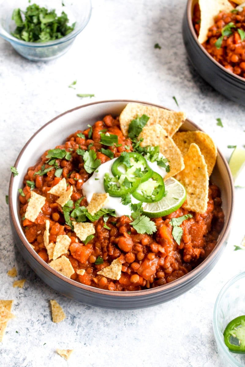 A bowl full of vegan bean & lentil chili topped with sour cream, cilantro, tortilla chips, and jalapeño.