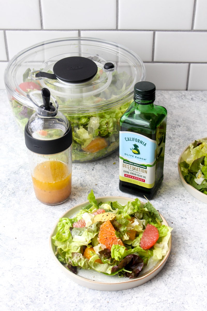 A plate with winter citrus avocado salad surrounded by a salad shaker, salad spinner, and olive oil.