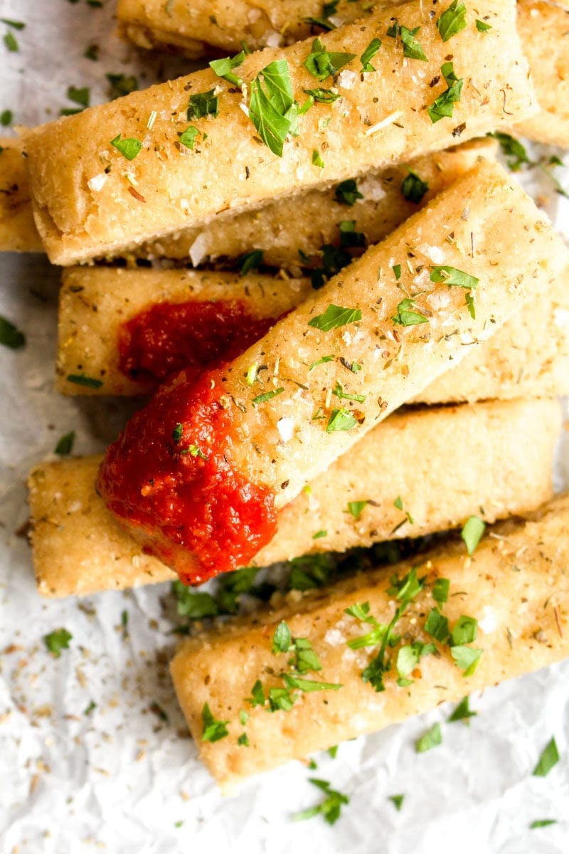 A pile of gluten free Italian herb breadsticks with marinara sauce.